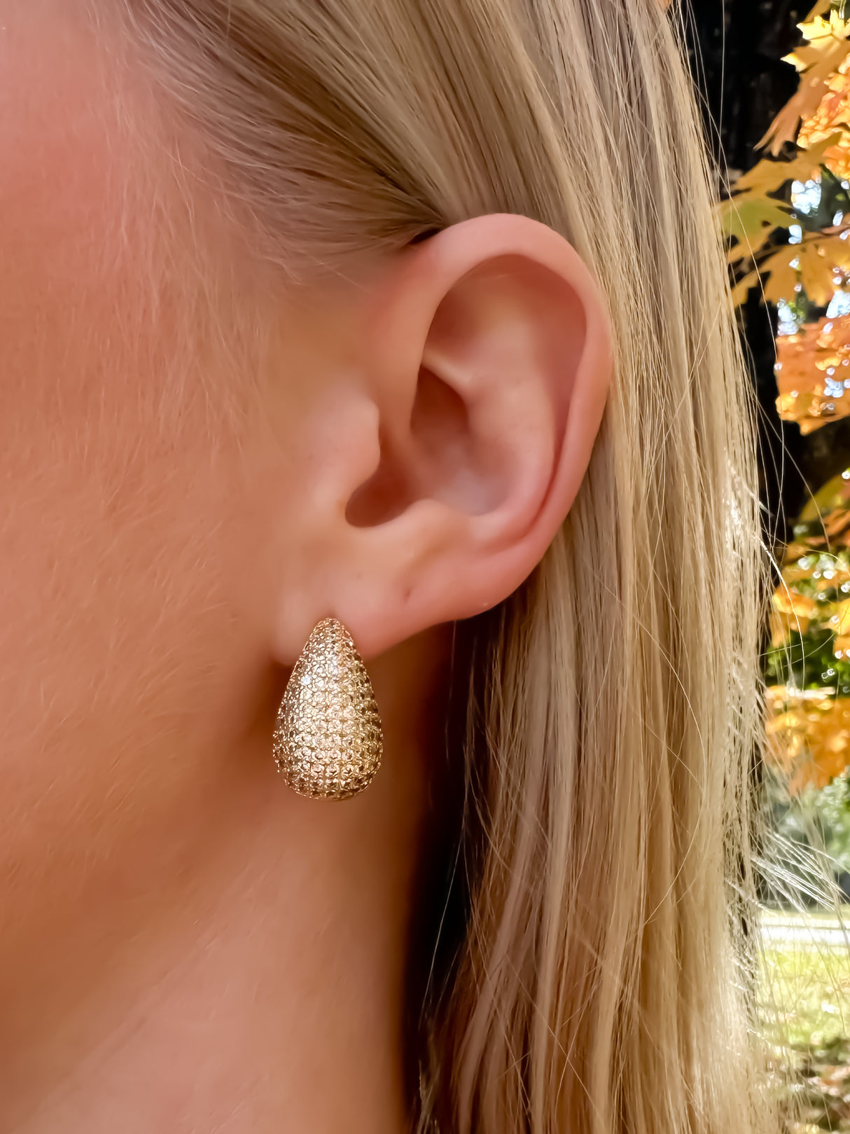 A close-up of Kinsey Designs' Alice Earrings, gold filled teardrop style, worn on a woman's ear with autumn leaves in the background.