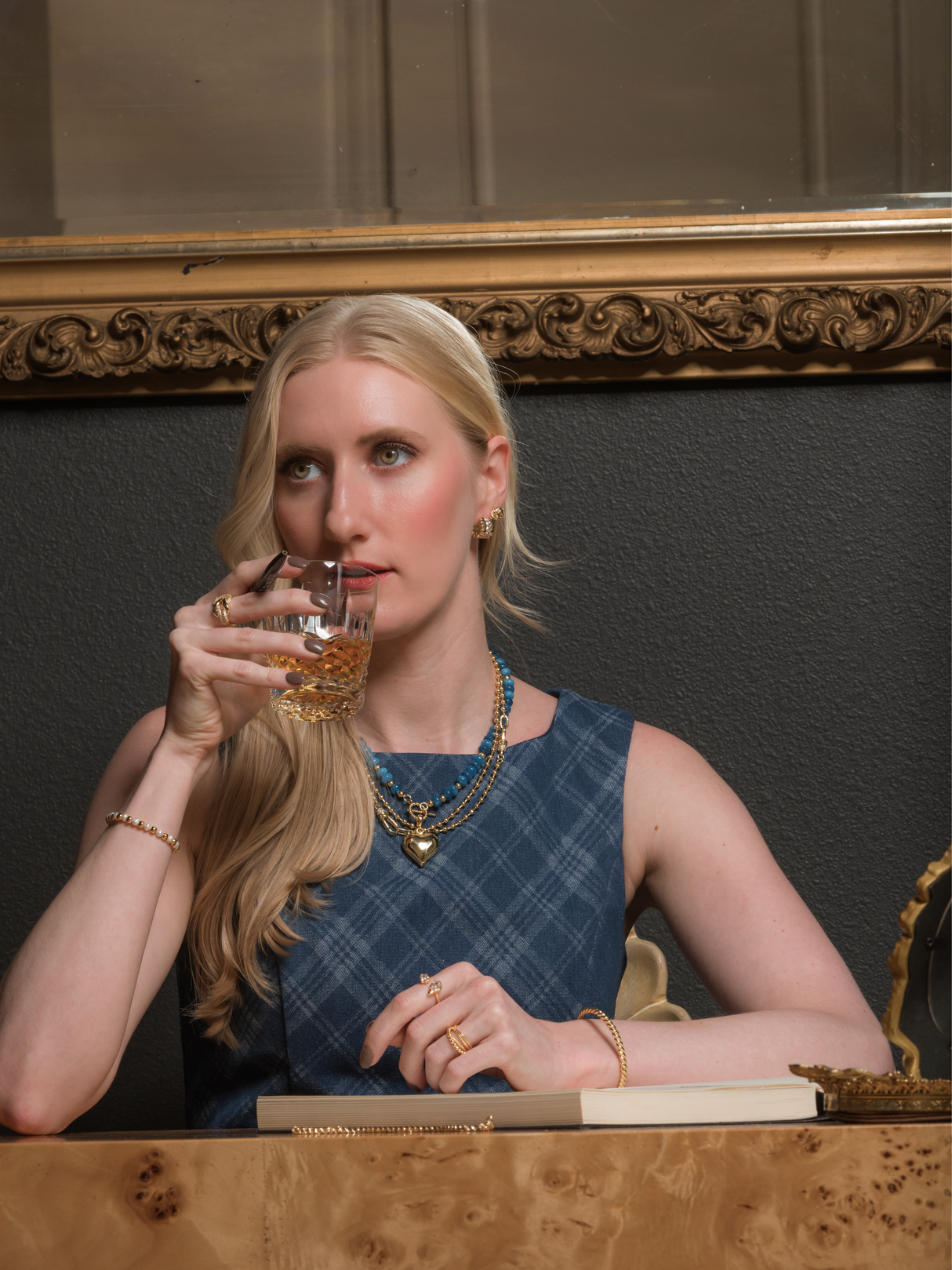 A woman in a blue dress, wearing the Ellis Huggie Earring by Kinsey Designs, sits at a table sipping from a glass, appearing thoughtful.