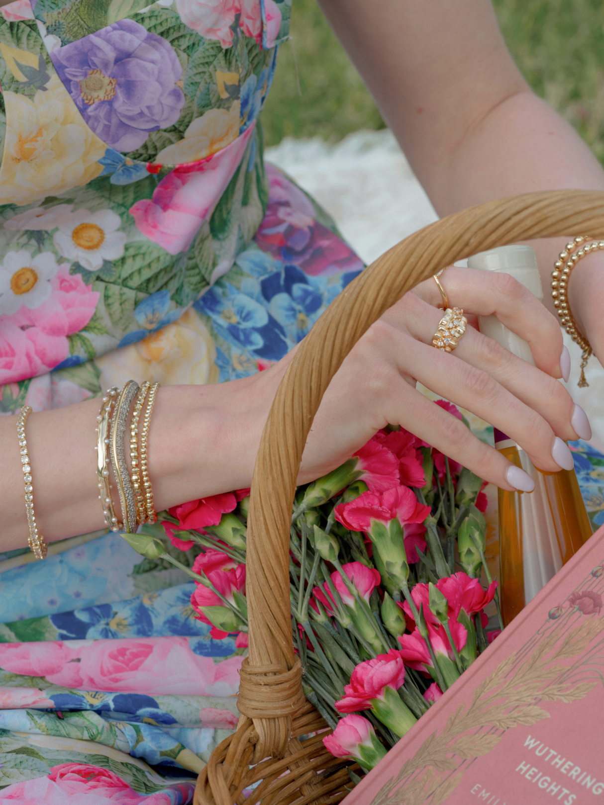A woman in a floral dress holds a basket with pink flowers, a book, and a bottle, accessorized with gold jewelry and the Kinsey Designs Graduating Bead Stretch Bracelet.