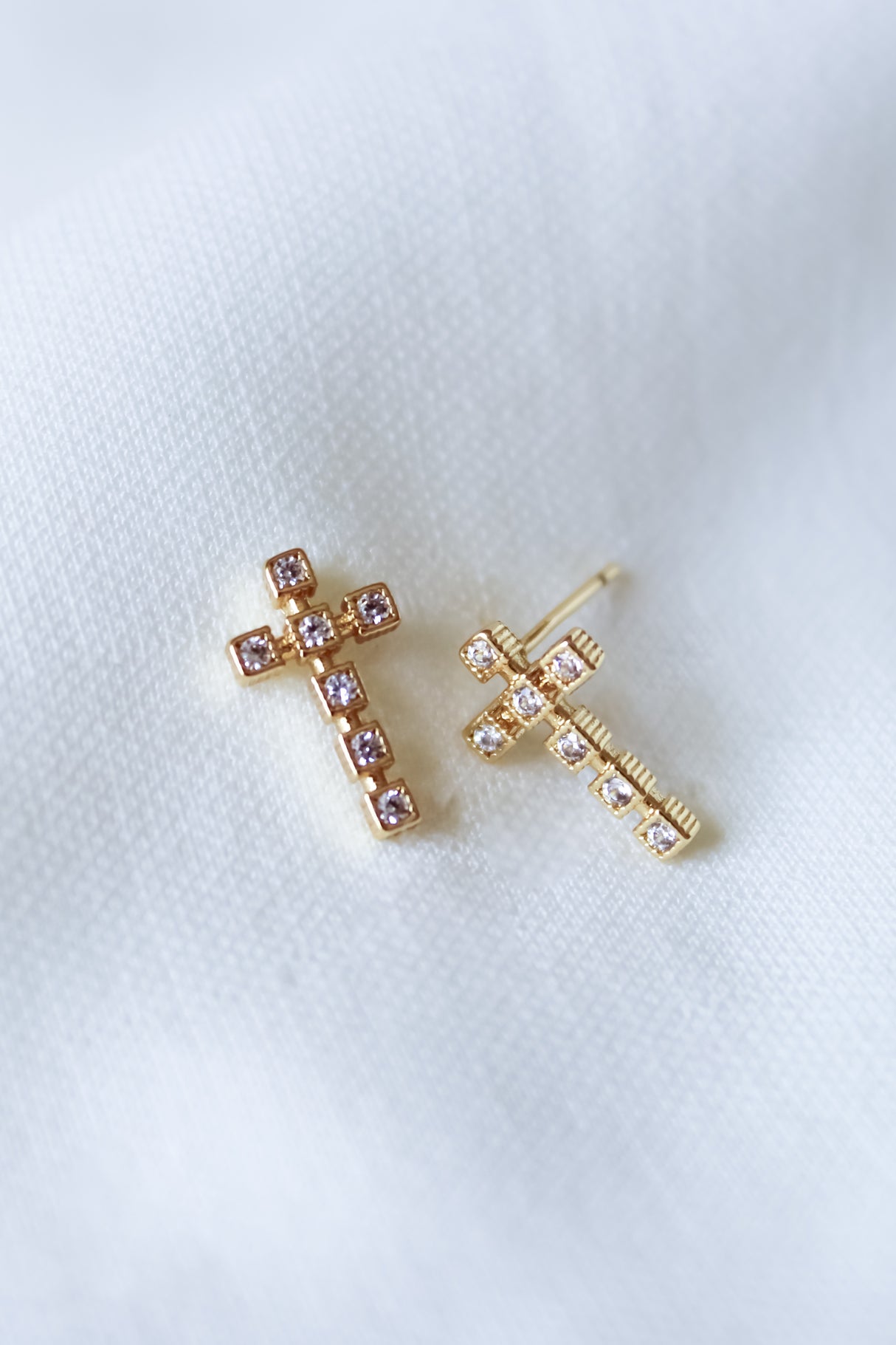 The Easton Stud by Kinsey Designs features gold filled cross-shaped earrings with small gems, displayed on soft white fabric. Ideal for a sample sale or final sale event.
