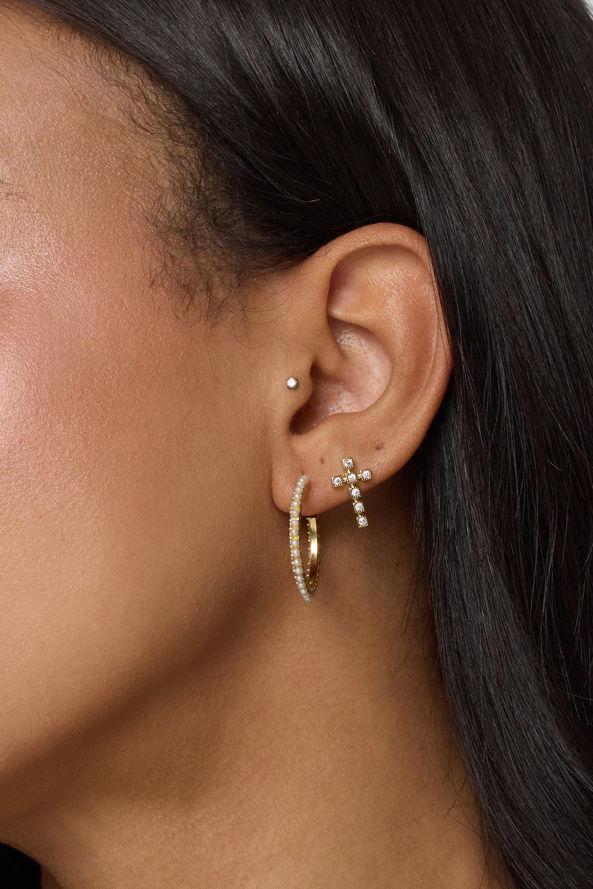 Close-up of a woman's ear with three earrings: the Easton Stud by Kinsey Designs, a cross, and gold filled hoops with tiny stones. Discover these perfect pieces at our sample sale.