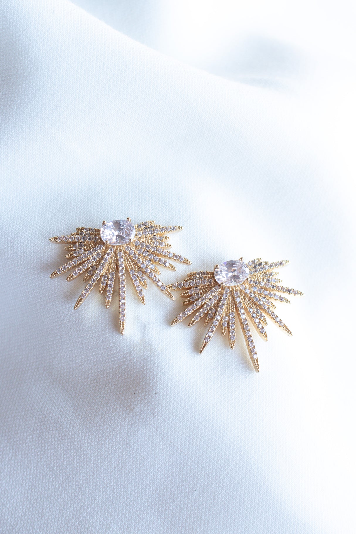 Kinsey Designs' Elaine Post gold-filled starburst earrings, adorned with sparkling cubic zirconia gemstones, showcased on a soft white fabric background.