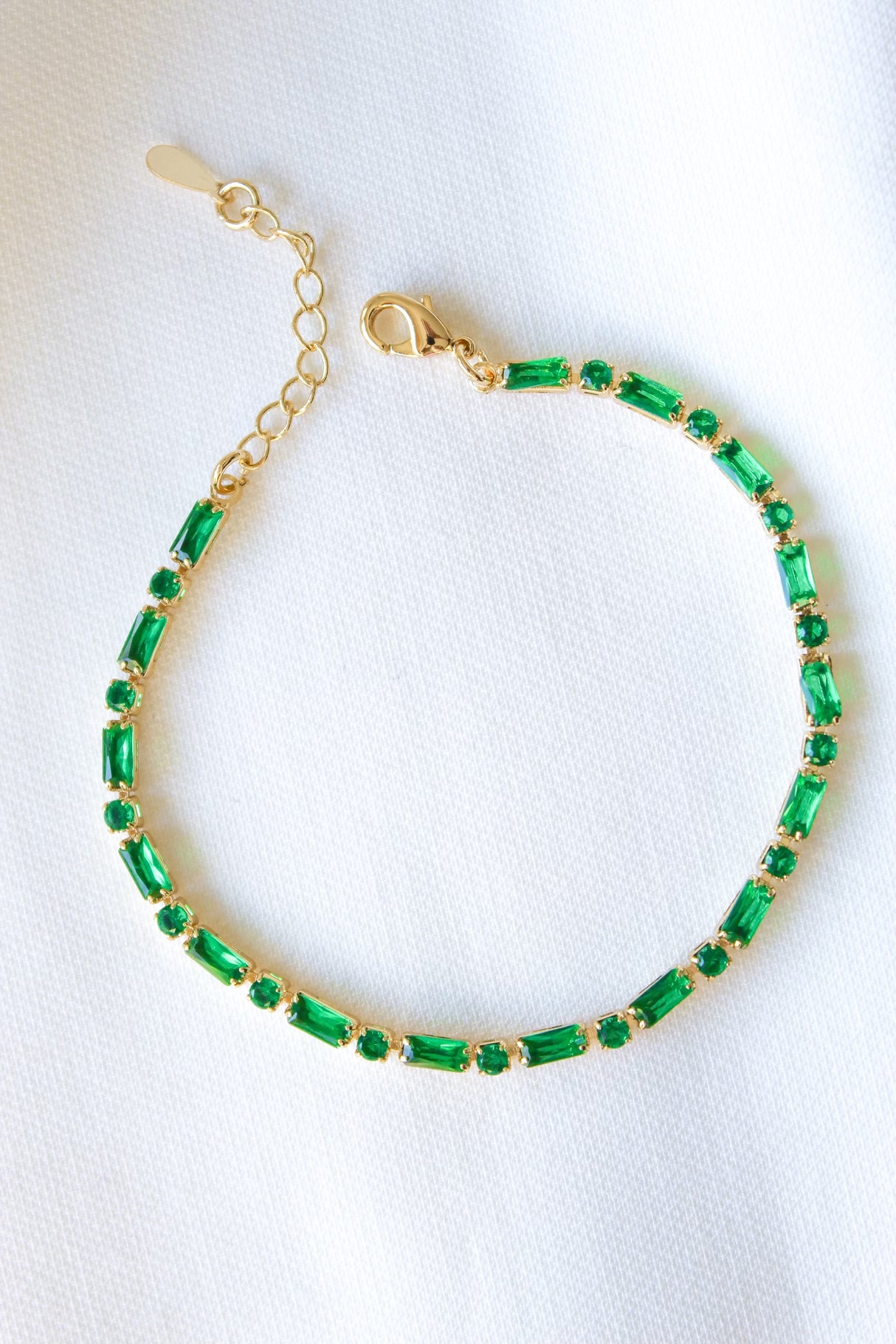 The Ella Bracelet by Kinsey Designs Archives features gold plating and rectangular green gemstones, shown on a white fabric. This adjustable piece adds everyday elegance to any look.