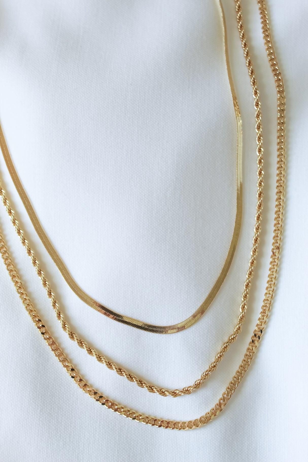 Four gold chains with unique patterns, including the Emile Layer Necklace by Kinsey Designs Archives, are displayed on a white fabric background.