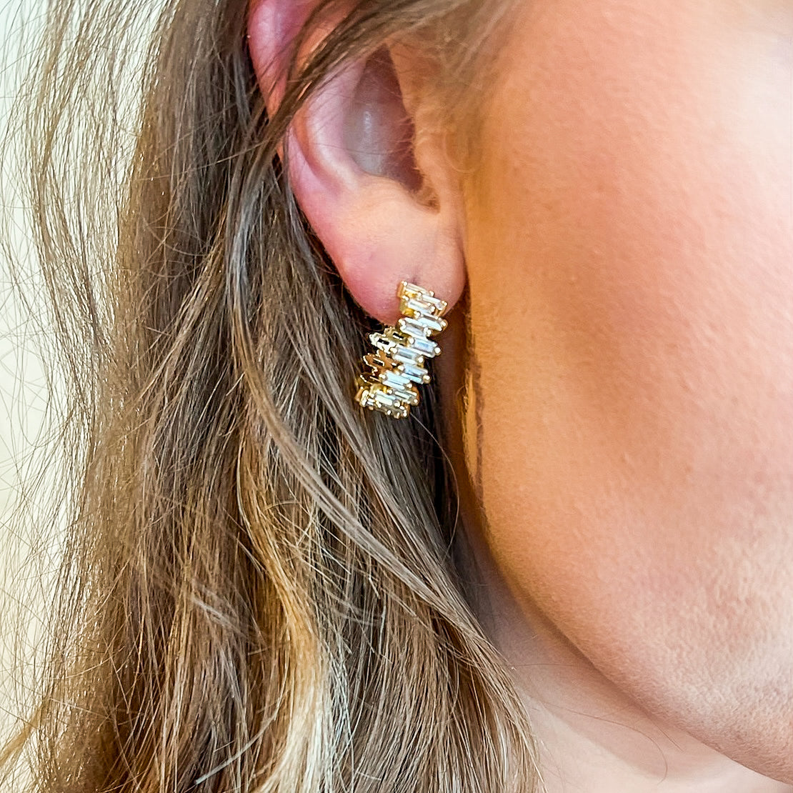 A close-up of a woman's ear wearing the Ettie Hoop by Kinsey Designs Archives, a 1" gold filled hoop earring featuring rectangular clear cubic zirconia gemstones.