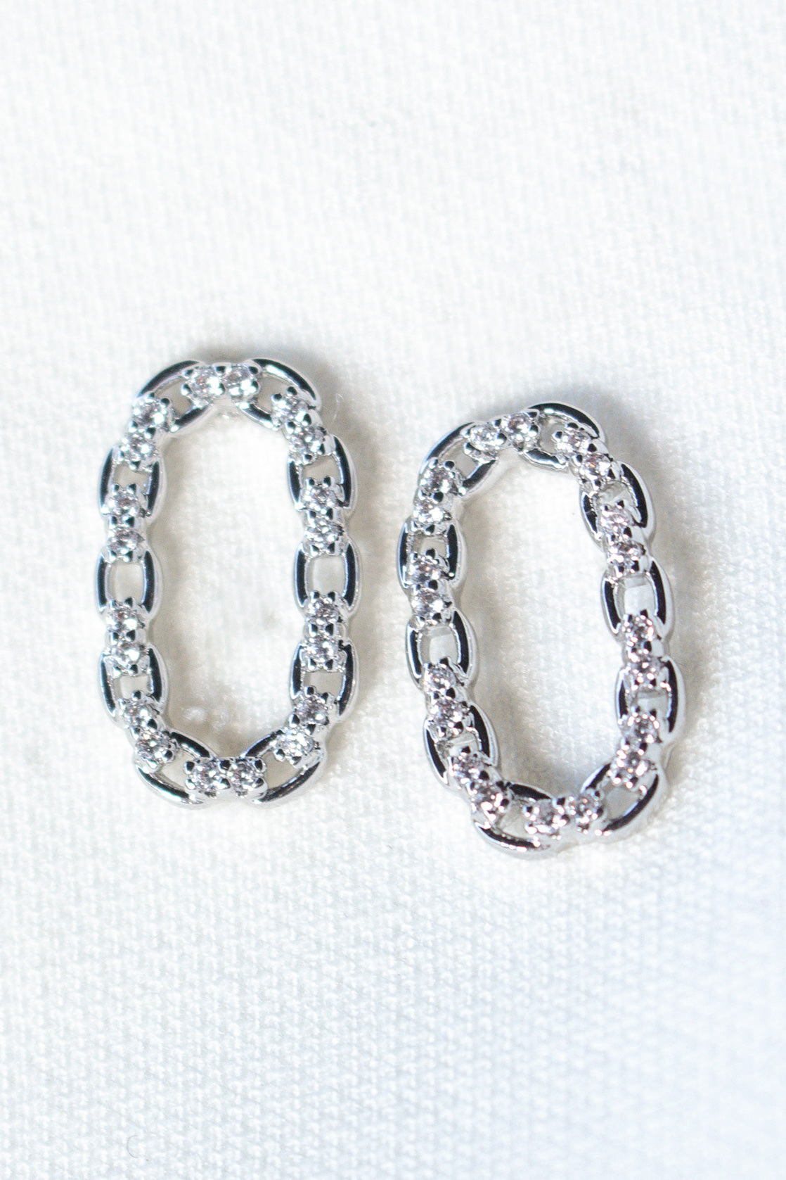 Everly Post Silver by Kinsey Designs—oval rhodium-plated brass chain earrings with clear cubic zirconia, displayed on a white textured surface. Perfect sample sale jewelry for those seeking elegant, sparkling style.