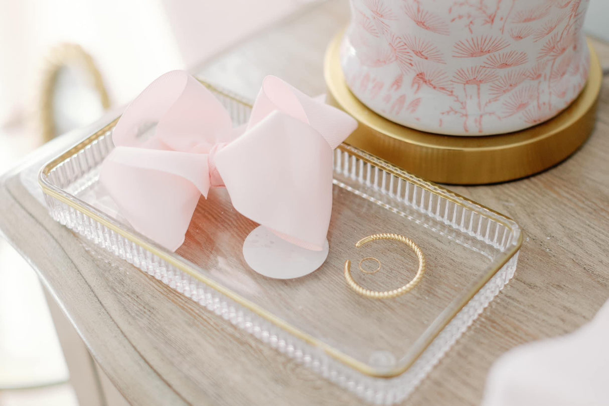 The Kinsey Designs Fallon Baby Cuff, paired with a pink bow and a gold ring, is displayed on a clear tray atop a wooden surface.