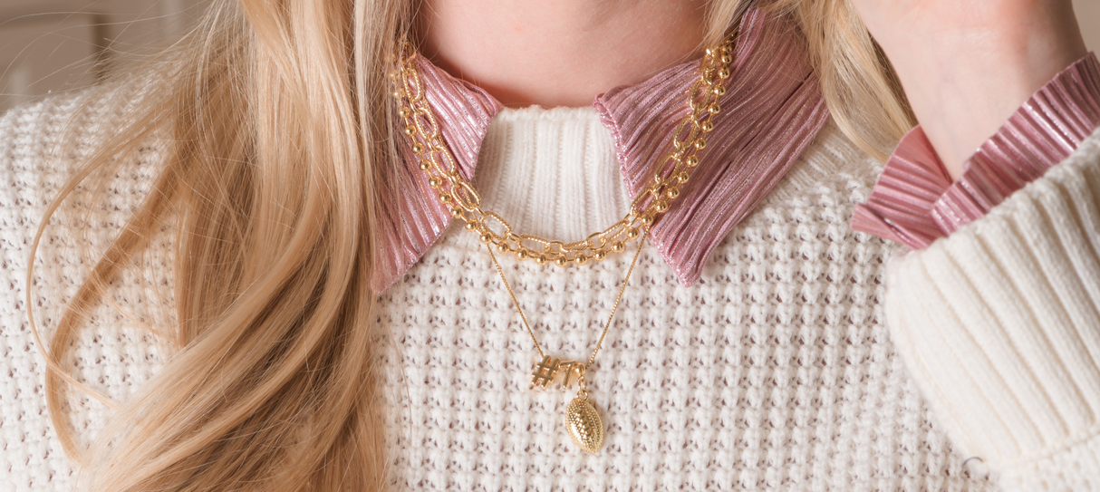 Smiling woman in a cap holding a glass, showing off game-day charm with Kinsey Designs’ Football Grip Charm necklace, paired with layered jewelry, a cream sweater, and a striped shirt.