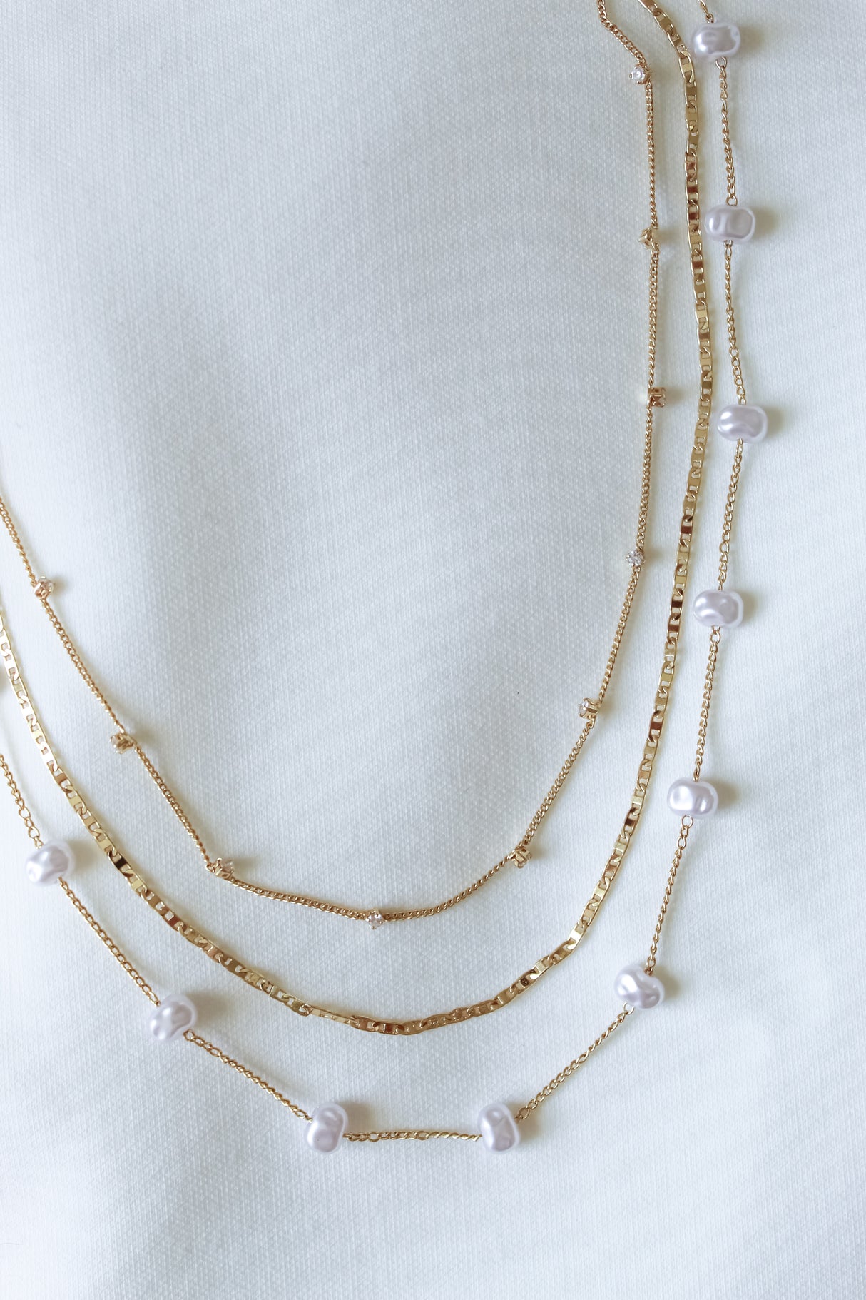 The Folly Layer Necklace by Kinsey Designs Archives features delicate gold chains and small pearls, displayed on a white textured fabric background.