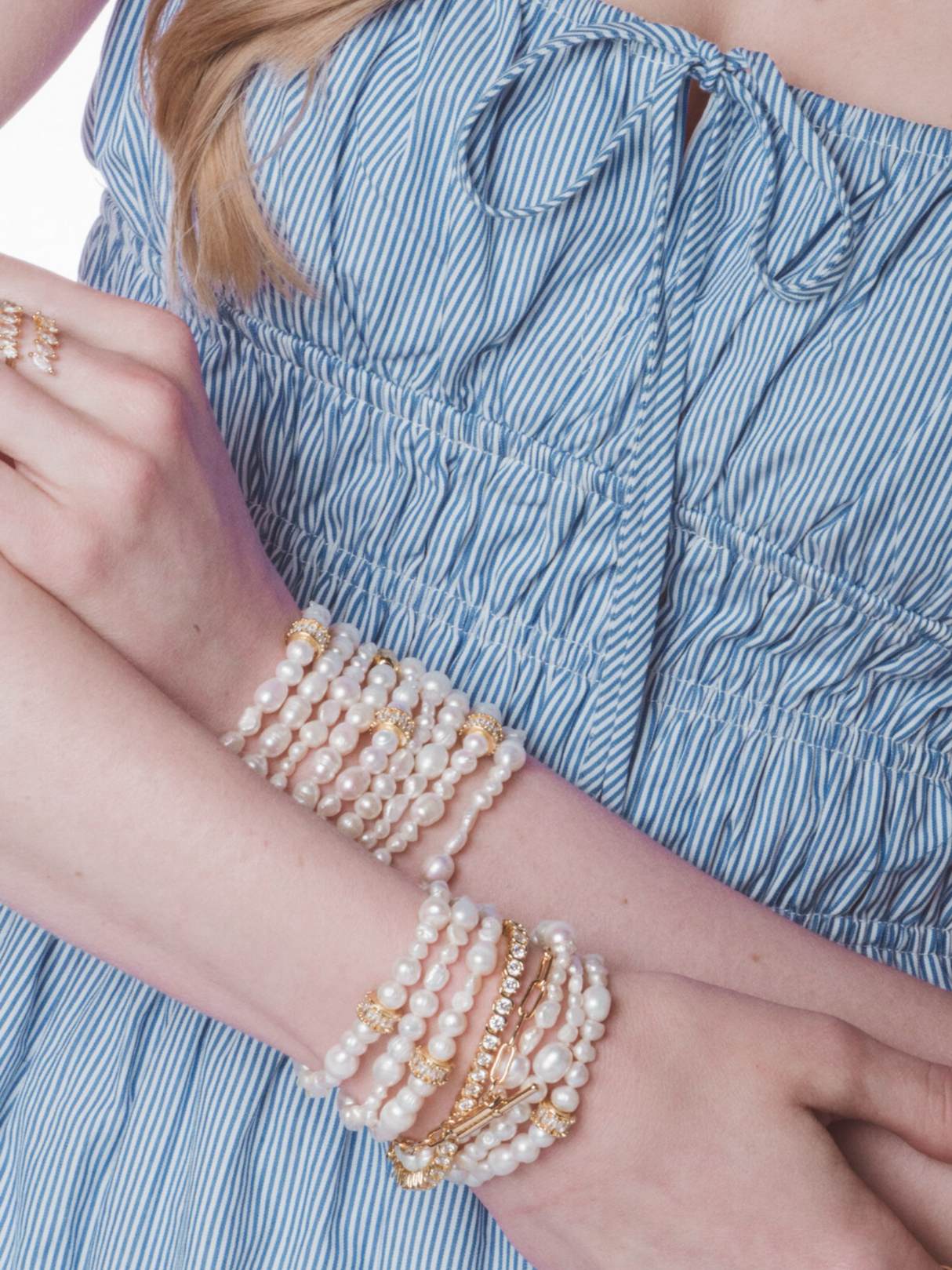 A woman in a blue striped dress wears multiple pearl and gold stretch bracelets on her wrists, including the stunning Kinsey Designs Freshwater Pearl Stretch Bracelet.