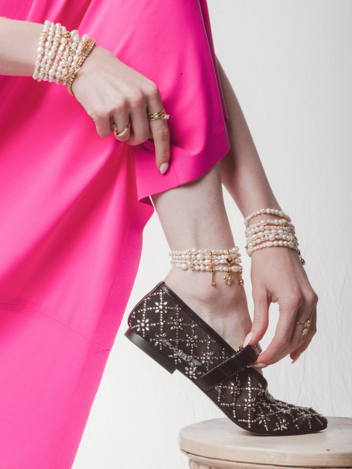 A woman in a bright pink dress, pearl jewelry, and black patterned shoes points to her ankle, showcasing the Kinsey Designs Freshwater Pearl Stretch Anklet.
