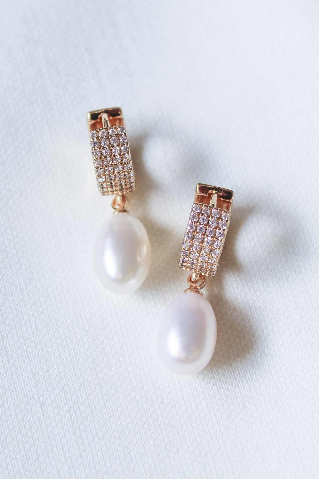 The Gemma Pearl Huggies by Kinsey Designs Archives feature gold-filled hoops with dangling white pearls, elegantly displayed on a white textured fabric background.