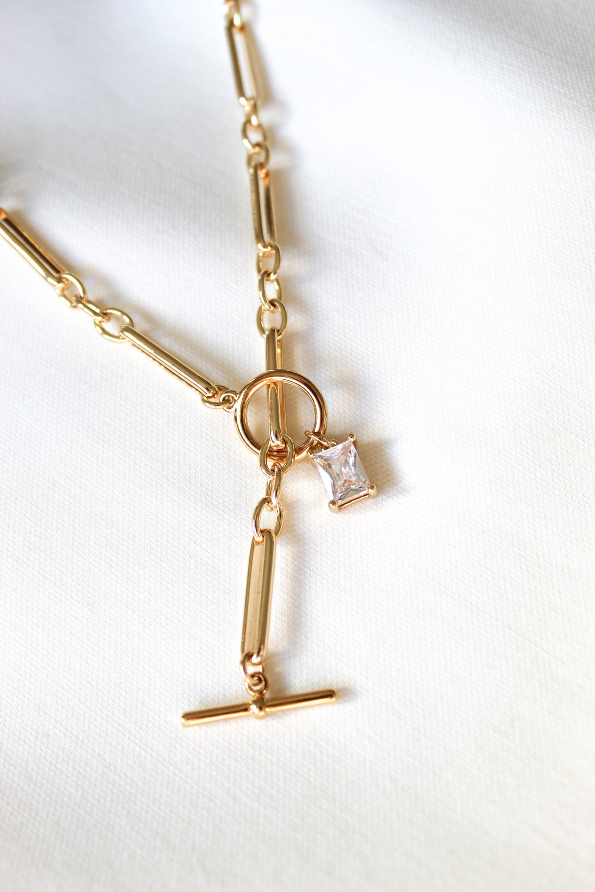 The Kinsey Designs Gilmore Necklace is a gold filled toggle pendant necklace with a rectangular clear gemstone, shown on a white fabric background.