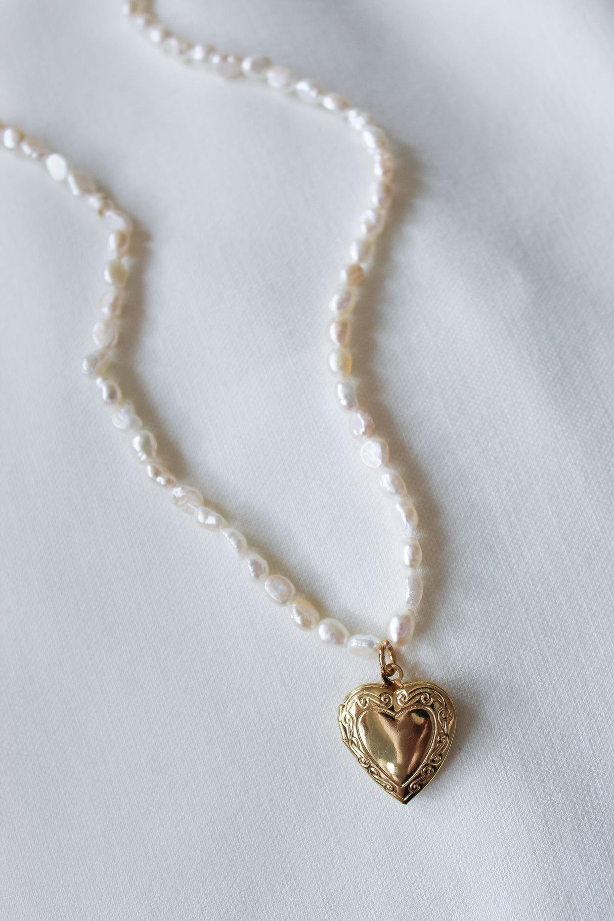 The Giselle Pearl Necklace by Kinsey Designs showcases lustrous freshwater pearls and a gold heart locket pendant, beautifully presented on a white fabric background.