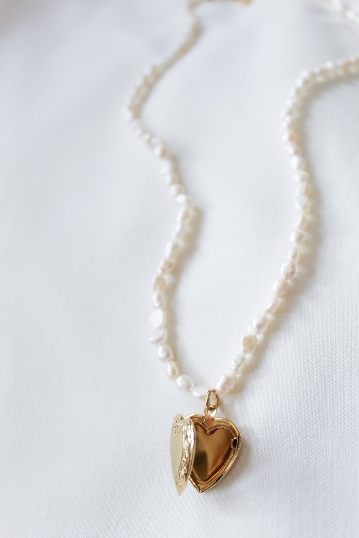 The Kinsey Designs Giselle Pearl Necklace, featuring freshwater pearls and an open gold heart locket, rests on a white fabric background.