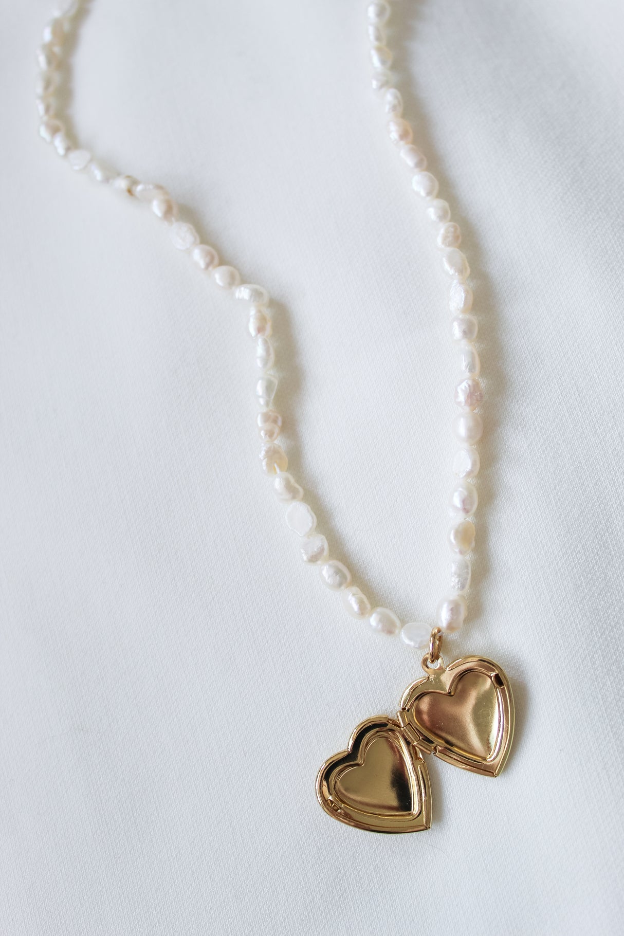The Giselle Pearl Necklace by Kinsey Designs showcases lustrous freshwater pearls and a delicate gold heart locket, elegantly displayed on a white fabric background.