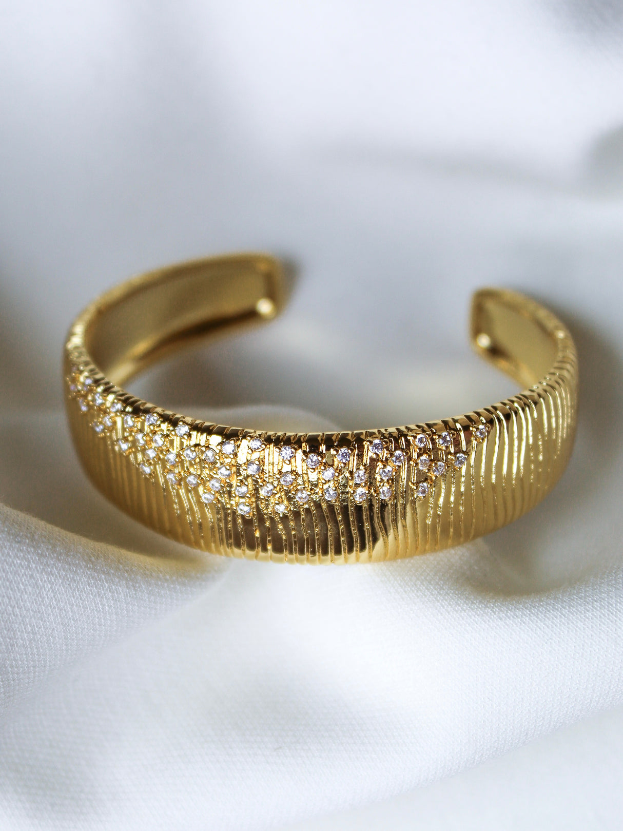 The Glinda Stardust Cuff Bracelet by Kinsey Designs features a gold-tone, textured surface with rows of sparkling CZ stones, elegantly displayed on white fabric.