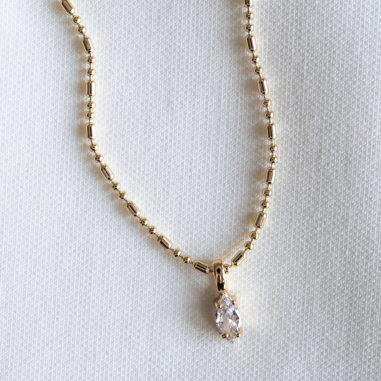 The Grace Drop Necklace by Kinsey Designs is a gold beaded statement piece with a marquise-cut clear gemstone pendant, shown on a white textured fabric background.