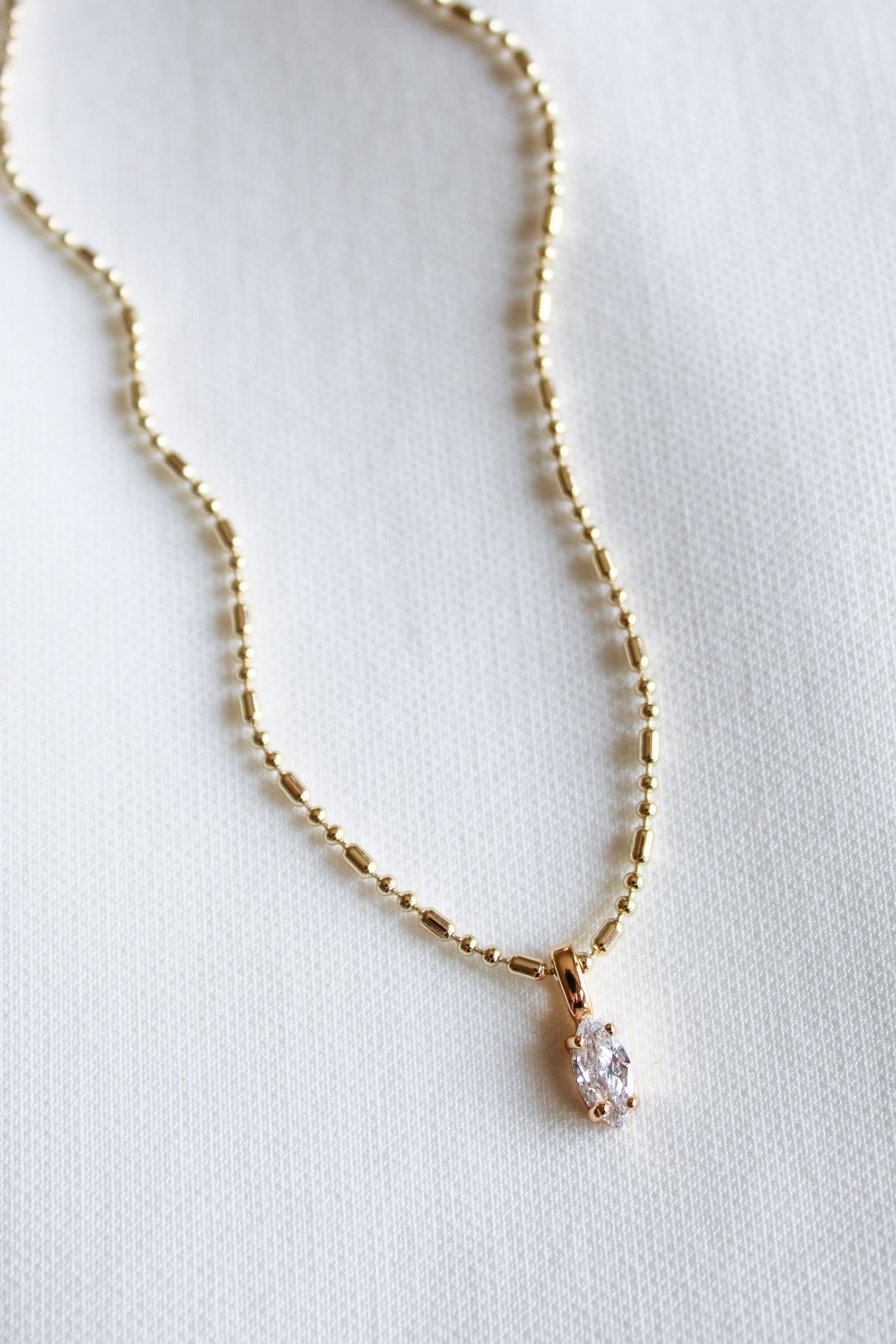 Kinsey Designs' Grace Drop Necklace features gold beads and a clear marquise-cut gemstone pendant, elegantly displayed on white fabric—a subtle statement piece ideal for any occasion.