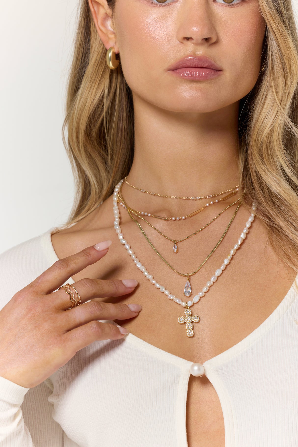 Woman wearing gold and pearl layered necklaces, featuring the Kinsey Designs Grace Drop Necklace with a cross pendant as a statement piece, along with gold hoops and a white top with pearl buttons.