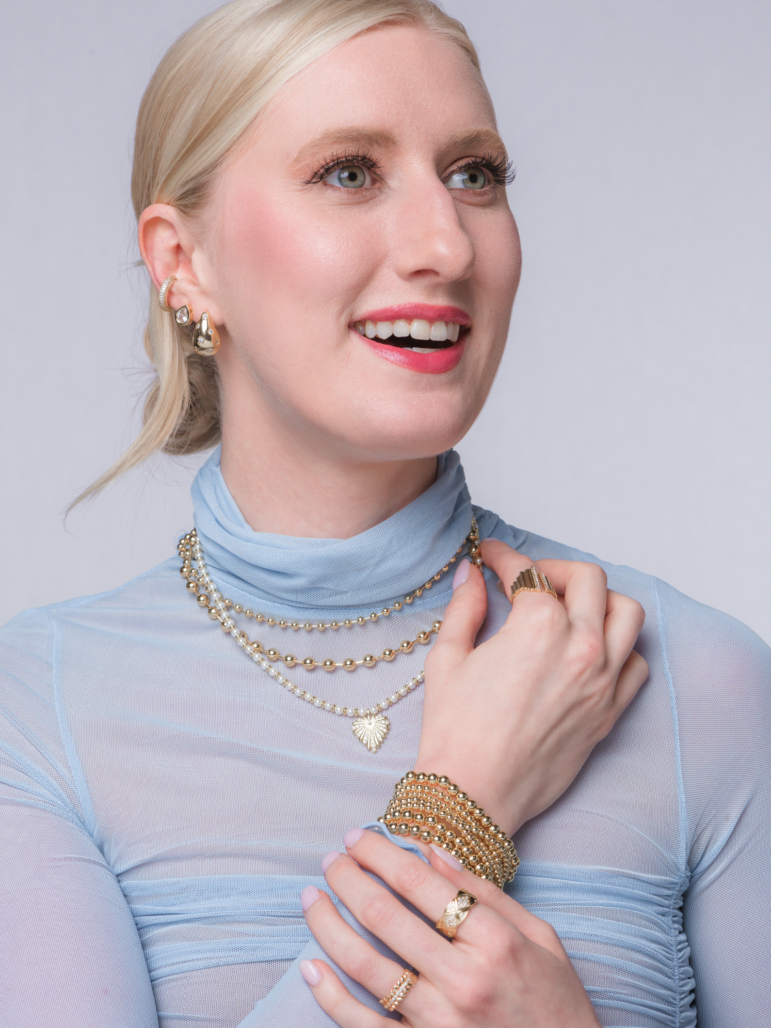 Smiling woman wears a sheer blue top with layered gold jewelry, featuring the Kinsey Designs Haven Necklace, a gold filled necklace, pearl beaded necklace, plus rings, earrings, and bracelets.