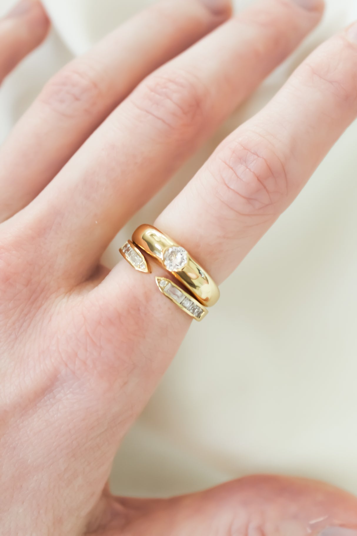 A hand wears two gold rings: the Kinsey Designs Hayden Ring with a central diamond, and a gold-filled ring adorned with small inset gemstones.
