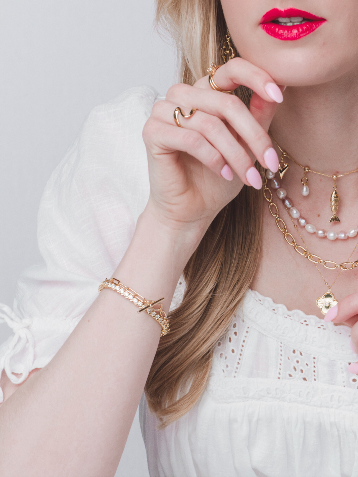 Woman in a white blouse wearing gold jewelry, the Kinsey Designs Holly Tennis Bracelet, pink nail polish, and bright red lipstick.