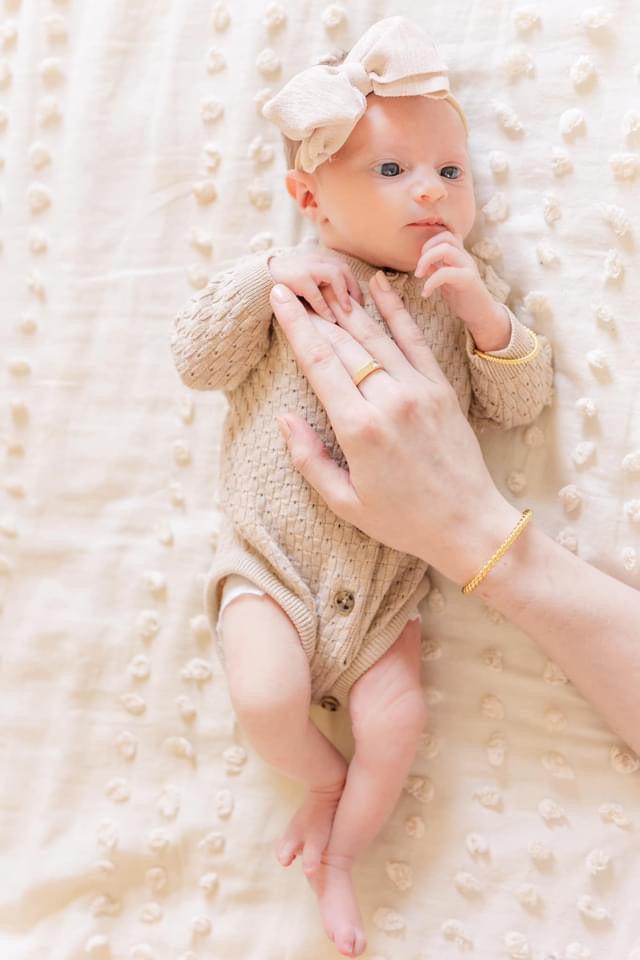 Newborn in a beige outfit with bow headband lies on a textured blanket, gently touching chest and wearing the adjustable, nickel-free Fallon Mama & Baby Cuff Set by Kinsey Designs.