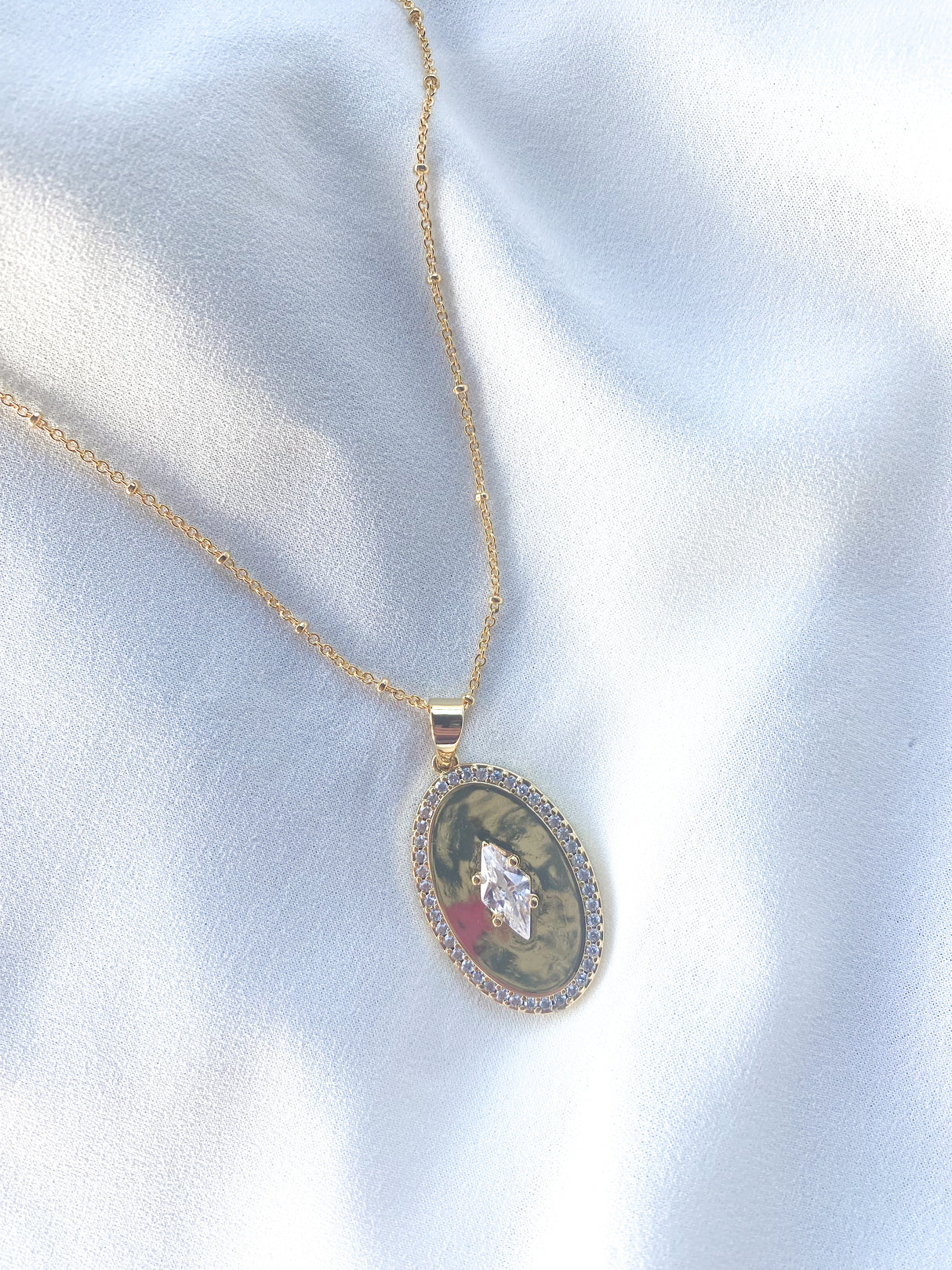 The Fique Necklace - Short by Kinsey Designs features an oval cubic zirconia and crystal detail, shown on white satin. Shop this stunning sample sale piece for an elegant touch to any look.