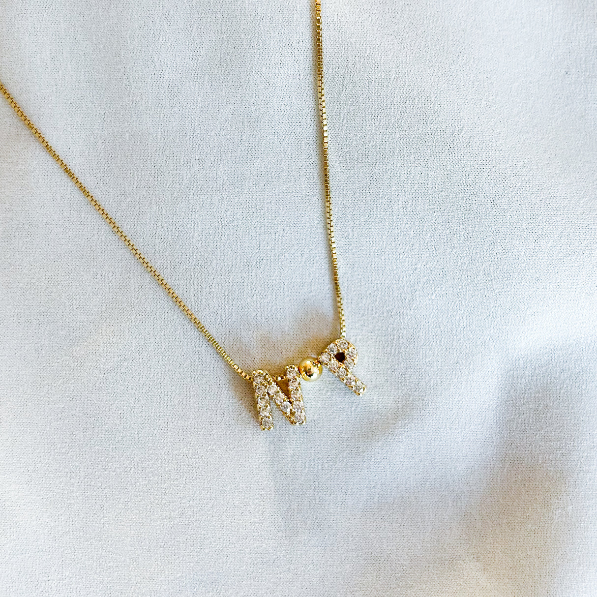 The Kinsey Designs Nurse Charm Necklace features gold filled "N" and "R" letter charms, perfect as a professional gift, and is beautifully displayed on a white fabric background.