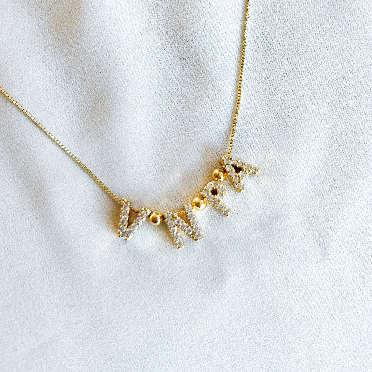 Gold filled Nurse Charm Necklace by Kinsey Designs featuring sparkling letter charms on a white fabric background—a chic and timeless piece for nurses.