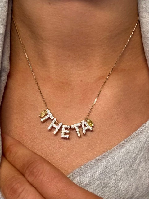 The Kinsey Designs THETA Charm Necklace features diamond-encrusted letters spelling "THETA," elegantly resting on the wearer’s neck.