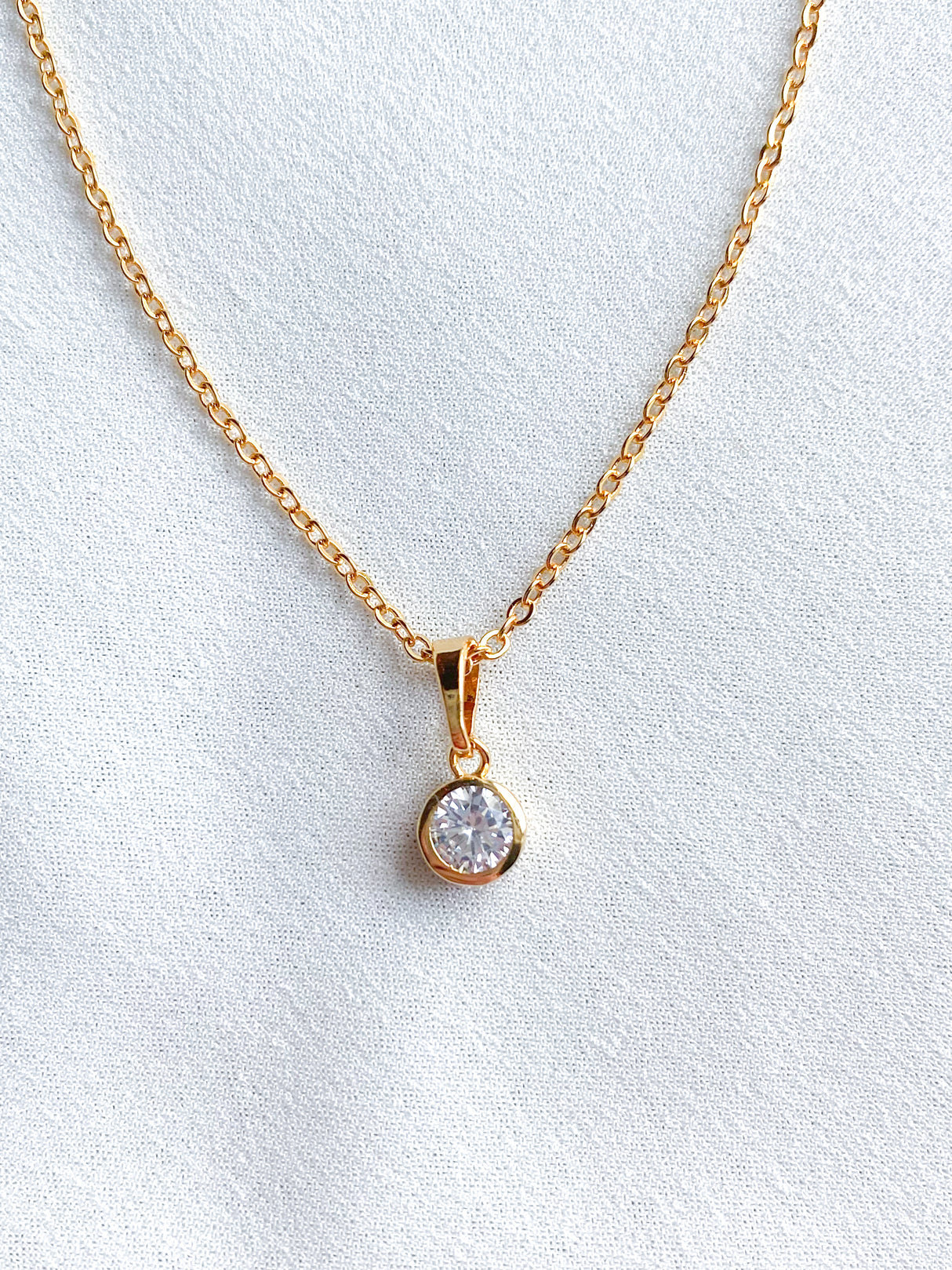 The Spritz Necklace by Kinsey Designs Archives features a round cubic zirconia pendant on an 18-inch gold filled chain, shown against a white fabric background.