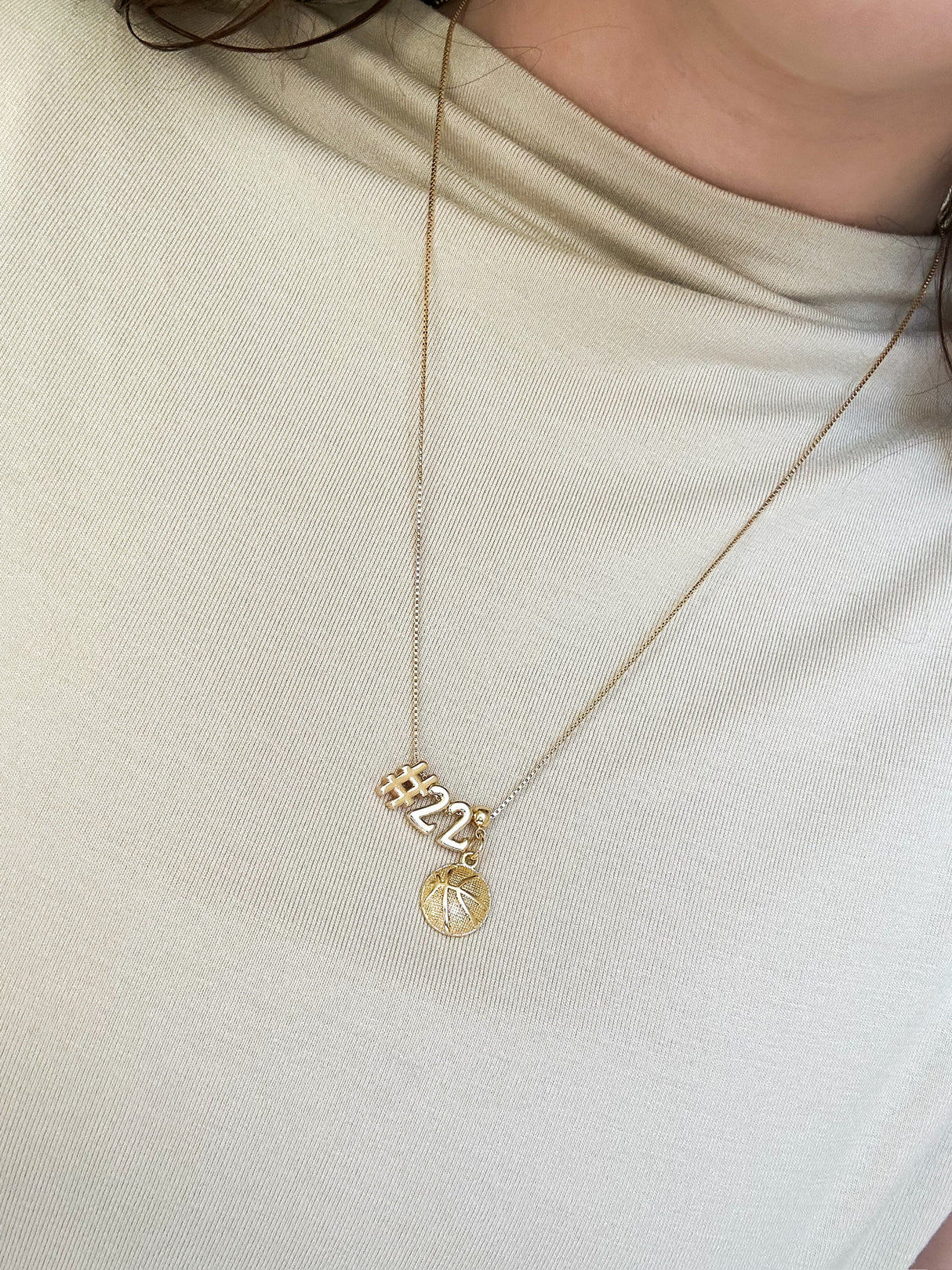 The Basketball Grip Charm necklace by Kinsey Designs, featuring a unique basketball charm and sunburst pendant, rests on a light beige top—ideal for fans of sporty, stylish jewelry.