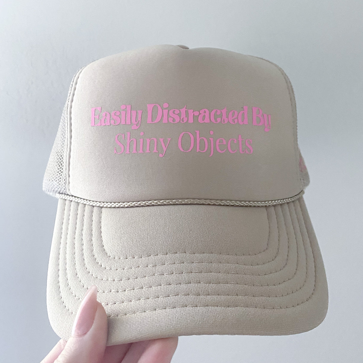 The Kinsey's Shiny Objects Hat by Kinsey Designs Archives is a beige trucker hat with pink "Easily Distracted By Shiny Objects" text, held in hand. Adjustable sizing ensures a comfy fit—an ideal summer accessory for lake days.
