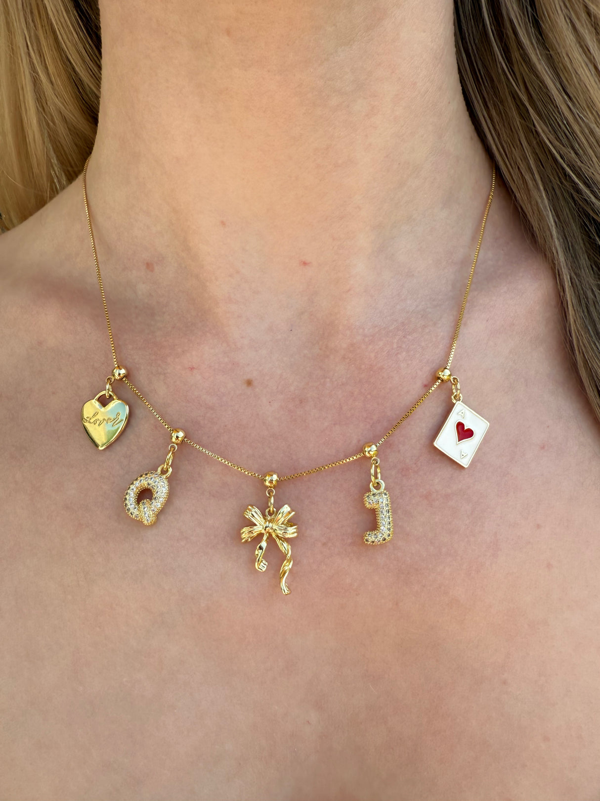 The Kinsey Designs gold necklace features five unique charms—heart, circle, bow, “J,” and the Ace Grip Charm—worn on a bare neck.
