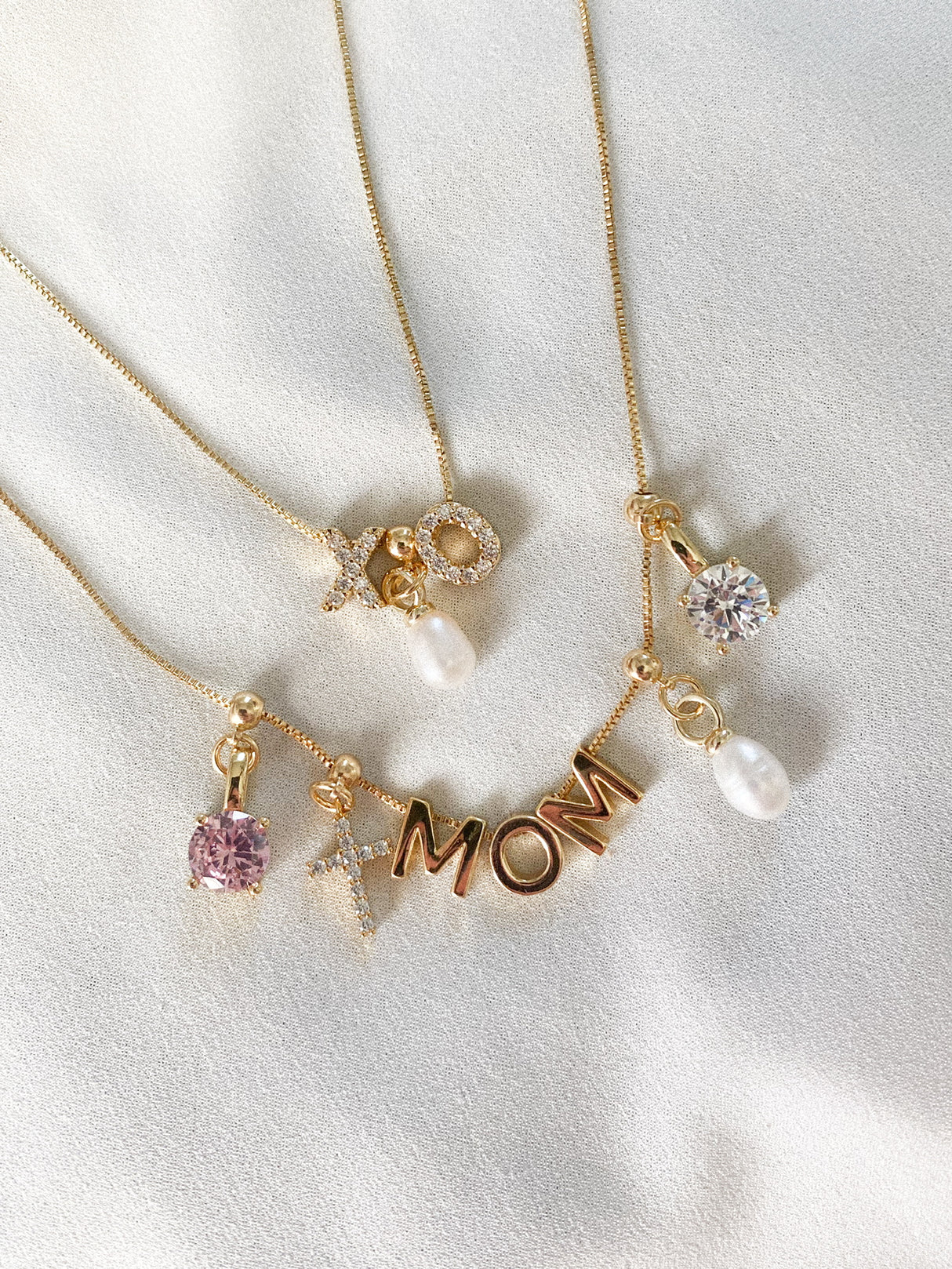 Three gold Kinsey Designs necklaces—charms reading "XO," "MOM," and "S"—feature pearls, gemstones, and the Cross Grip Charm crystal cross, arranged on white fabric.