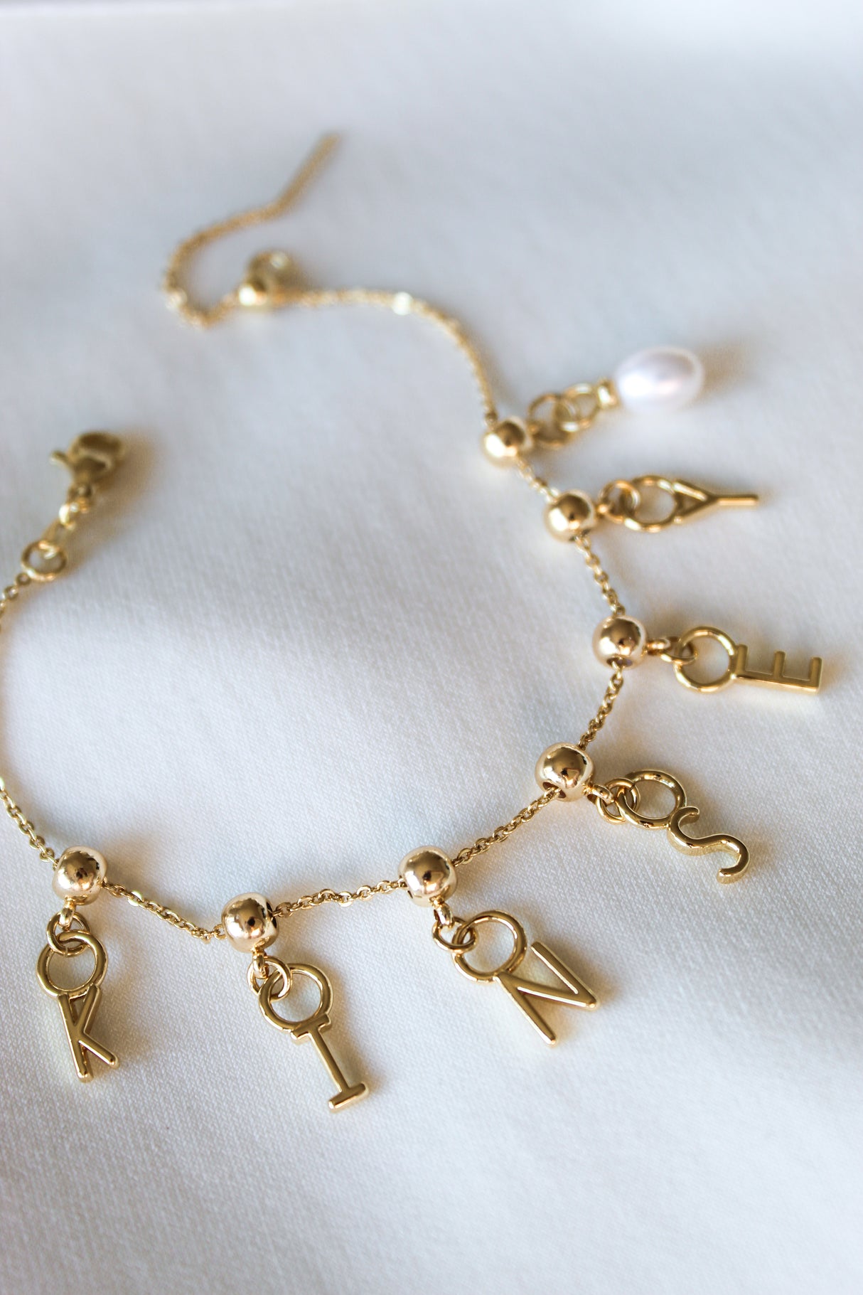 The Kinsey Designs Archives Letter Grip Charm gold bracelet features gold-filled charms spelling "KINDNESS" and a single white pearl, displayed on a white fabric background—a personalized jewelry piece celebrating positivity and elegance.