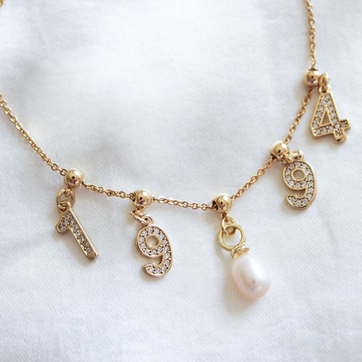 The Kinsey Designs Charm Bracelet Dainty Chain features number charms "1", "9", "9", "4" and a pearl, laid on white fabric—perfect for pairing with your favorite dainty chain or gold-filled bracelet for a chic layered style.