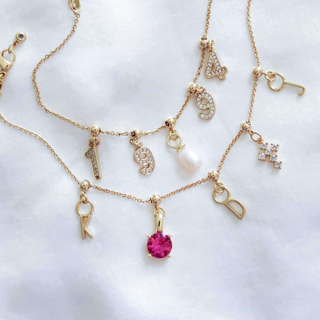 Kinsey Designs’ gold necklaces with letter and number charms, a pink gem, pearl, rhinestone details, and a Silver Birthstone Grip Charm are displayed on a white fabric background.