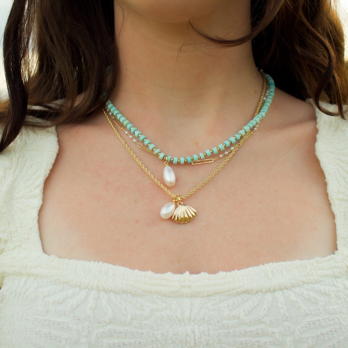 A woman wears a white textured top and layered necklaces, including a turquoise and gold necklace and the Kinsey Designs Archives Sailor Pearl Necklace featuring shell and teardrop pearl pendants.