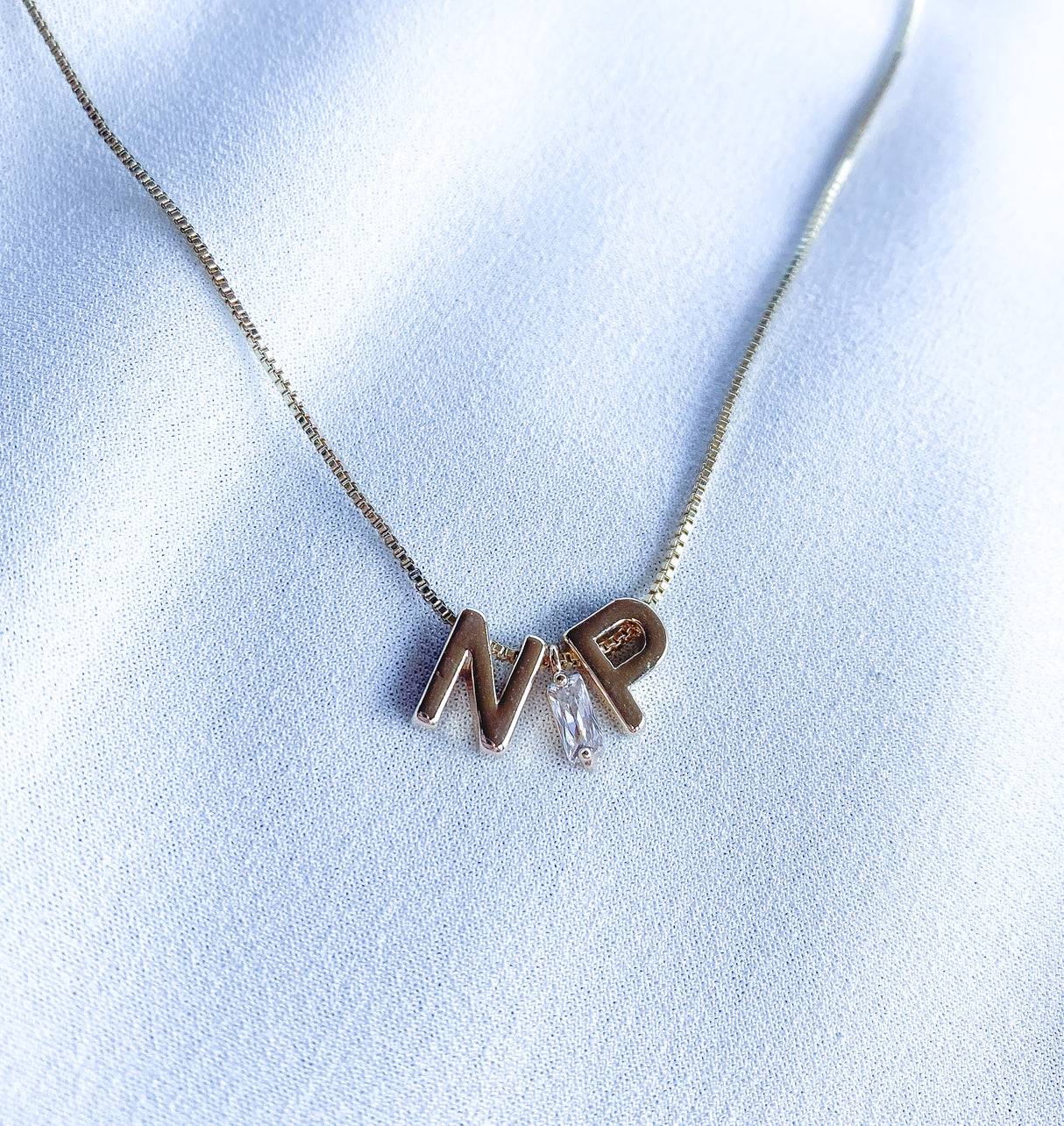 The Kinsey NP Adjustable Necklace by Kinsey Designs features "N" and "P" letter charms with a small crystal, perfect as a profession necklace, and is displayed on white fabric.