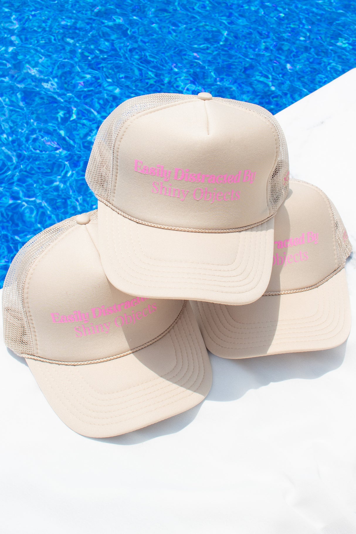 Three Kinsey Designs Archives "Kinsey's Shiny Objects Hat" in beige mesh with pink text, shown poolside—an ideal summer accessory with adjustable sizing for a perfect fit.