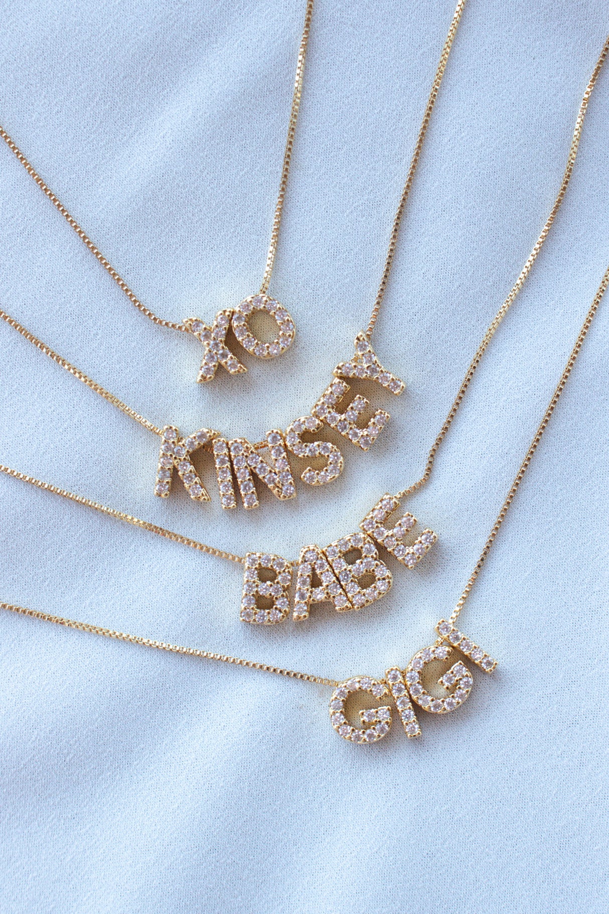 Four gold necklaces from Kinsey Designs, each with a LUX Letter Slide Charm—XO, KINSEY, BABE, and GIGI—rest on white fabric, featuring sparkling crystal-studded pendants for a personalized touch.