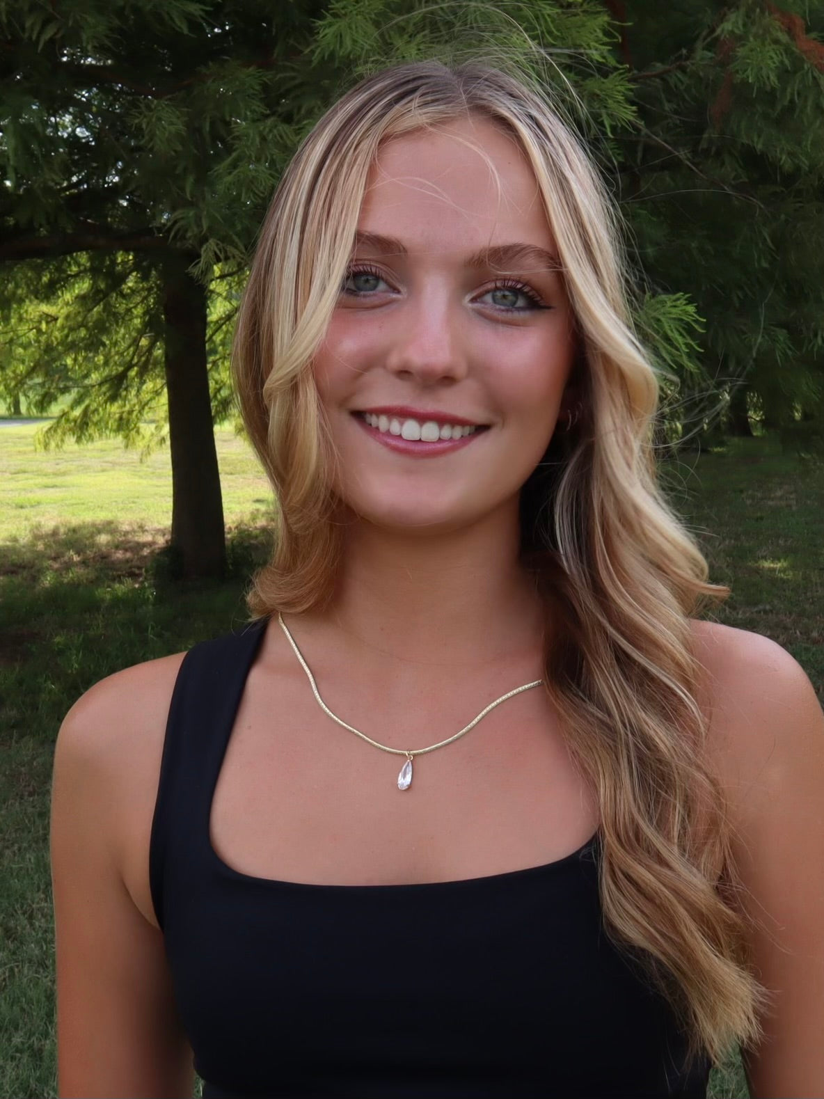 A young woman with wavy blonde hair smiles outdoors, wearing a black top and the Kinsey Designs Charlotte Drop Necklace.