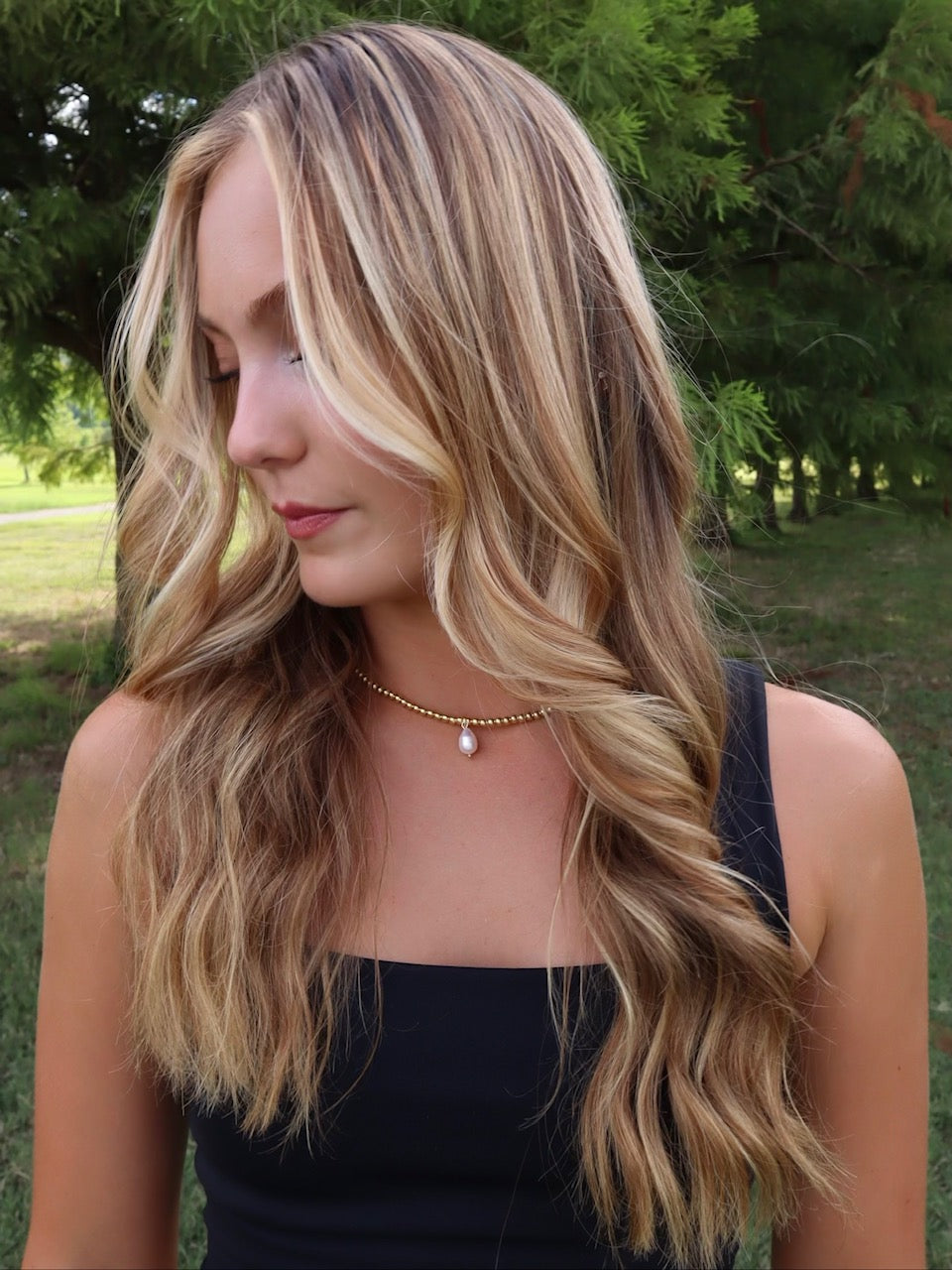 A young woman with wavy blonde hair stands outdoors before green trees, wearing a black top and the Reed Necklace by Kinsey Designs—a delicate pearl accessory.
