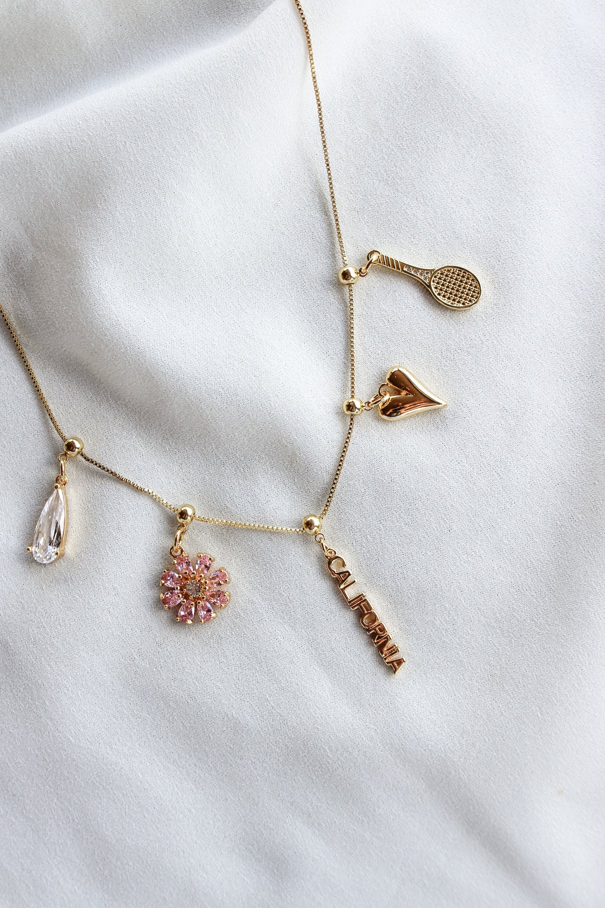 The Kinsey Designs Mississippi Grip Charm adds state pride to any necklace, featuring a gold bar with teardrop, pink flower, "California," sneaker, tennis racket, and is perfect for jewelry lovers seeking unique charm designs.