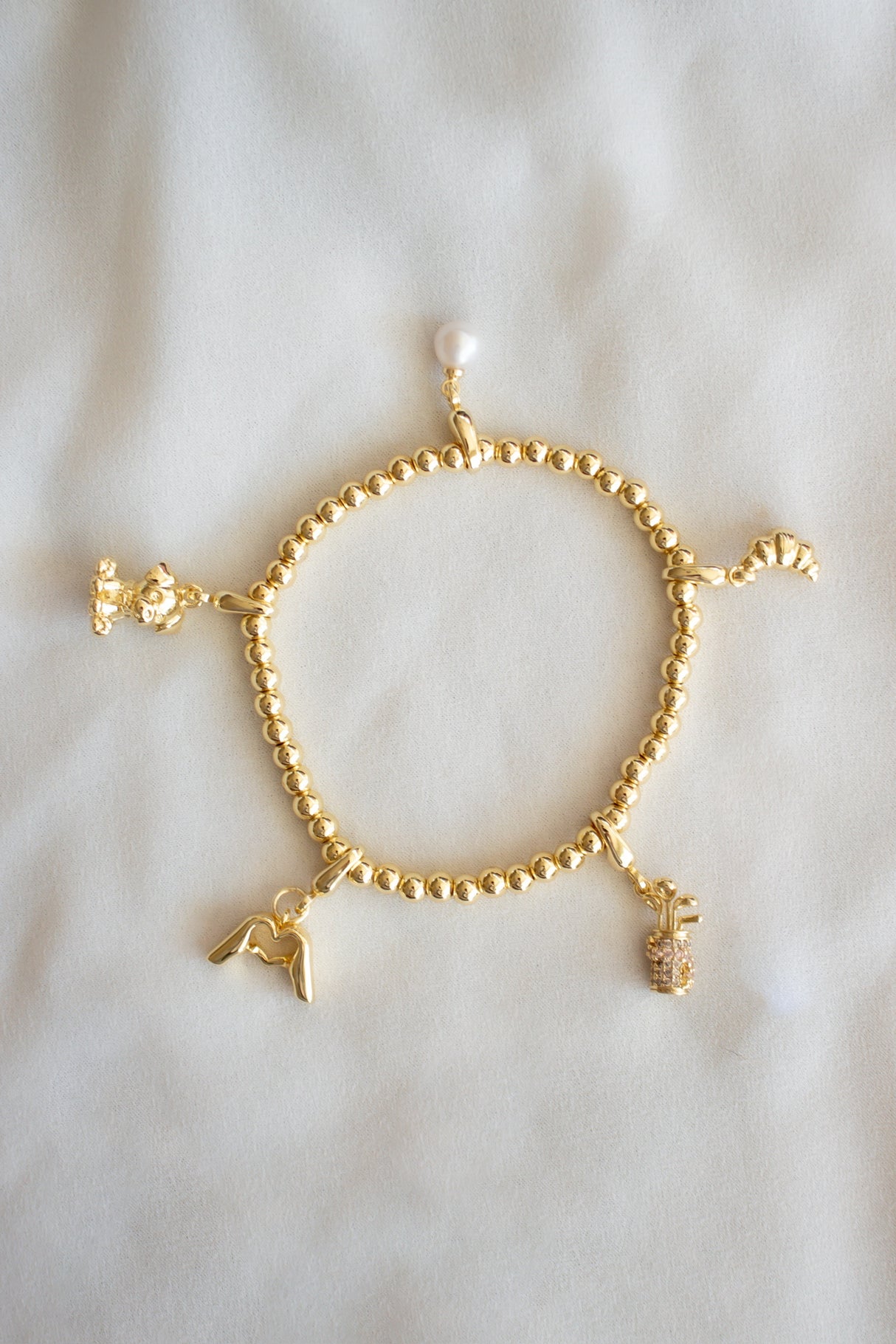 The Croissant Clip Charm by Kinsey Designs is featured on a gold filled beaded bracelet with four unique charm slides, shown on a soft, light-colored fabric background.