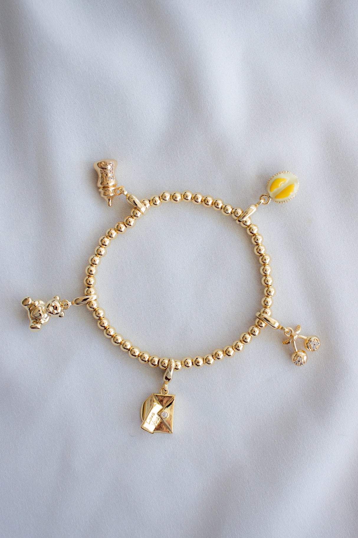 The Kinsey Designs Baby Bottle Clip Charm is styled on a gold filled beaded bracelet with four other unique charms from the Kinsey Charm Bar, displayed on a soft white fabric background.