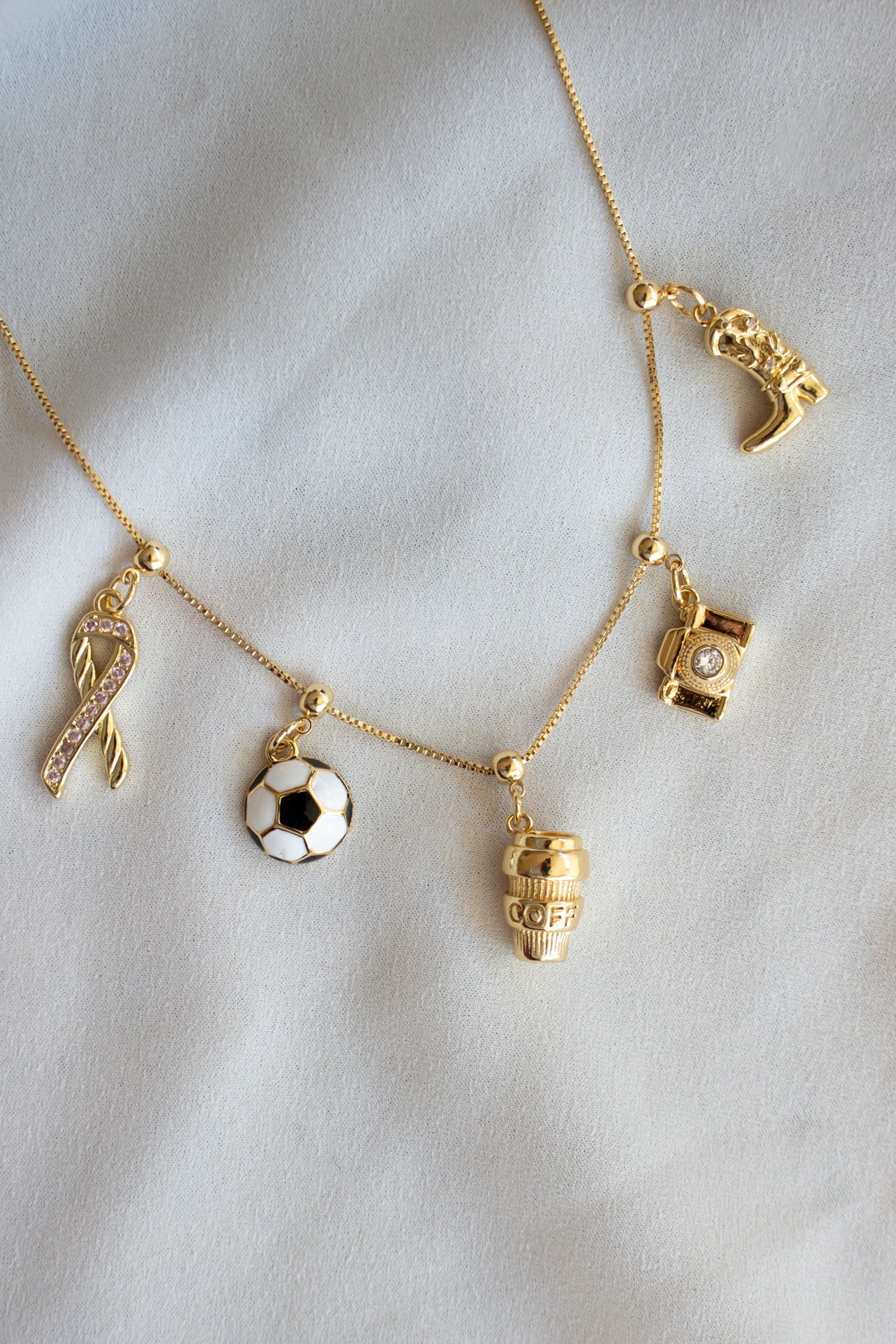 The Kinsey Designs Hope Grip Charm necklace features gold-filled dangling charms—a breast cancer awareness ribbon, soccer ball, coffee cup, camera, and soccer shoe—elegantly displayed on white fabric.
